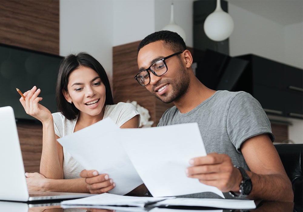 BIPOC-couple-sitting-together-happily-looking-at-laptop-and-paperwork