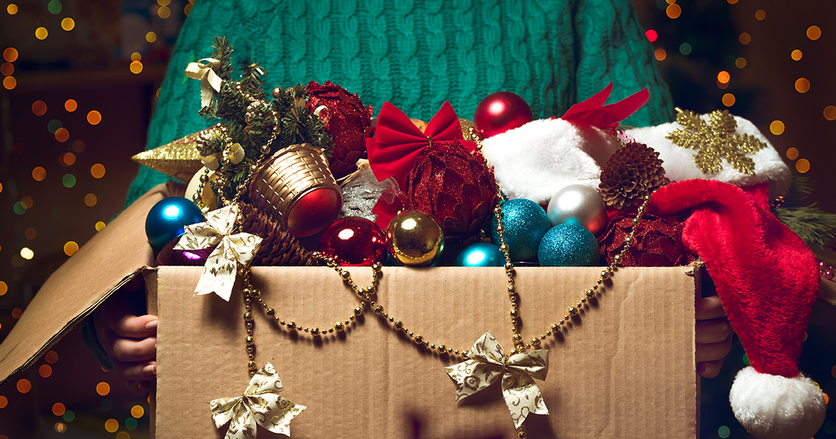 person wearing green sweater holding a box full of red, gold, and teal colored Christmas decorations