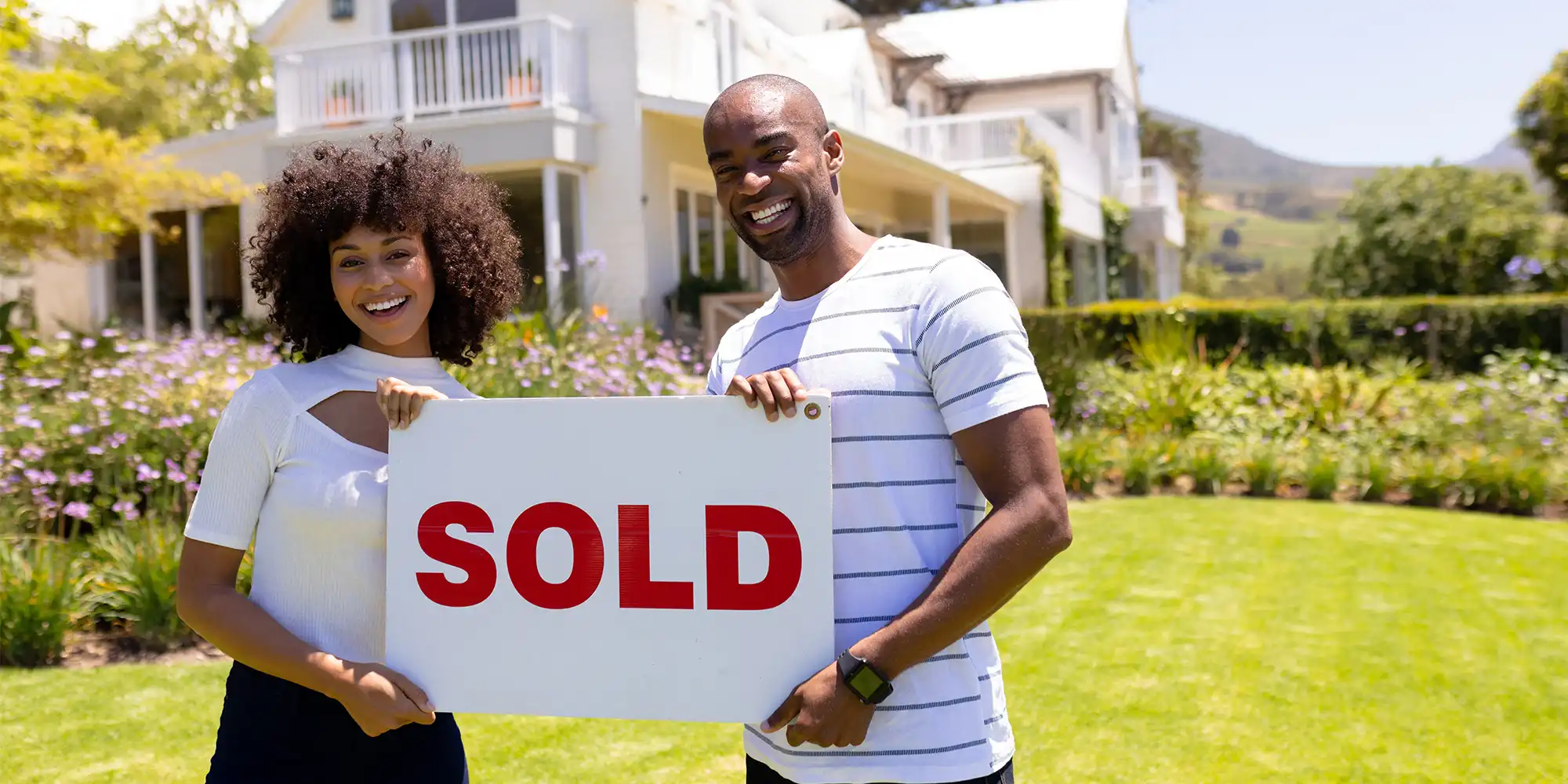 young african american couple standing outside a two story white colored home with a pretty garden holding a sign that says 
