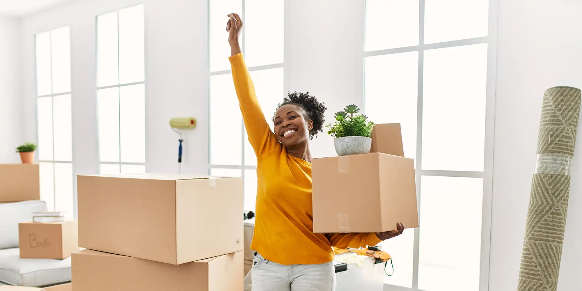 african american woman with hair pulled back wearing golden yellow shirt and light jeans standing with arm in the air in white livingroom with moving boxes