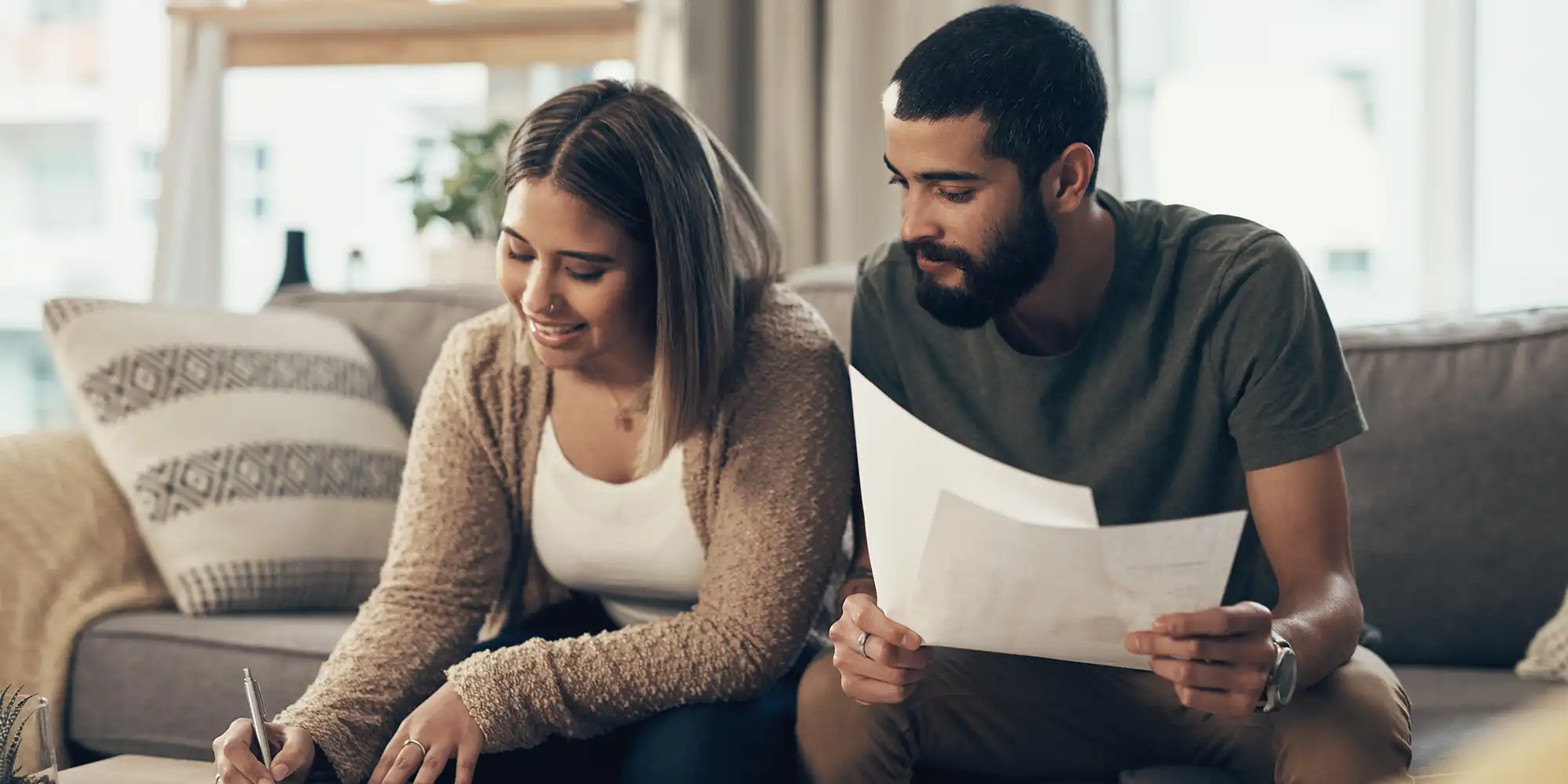young heterosexual BIPOC couple signing loan documents on a coffee table while sitting on brown sofa