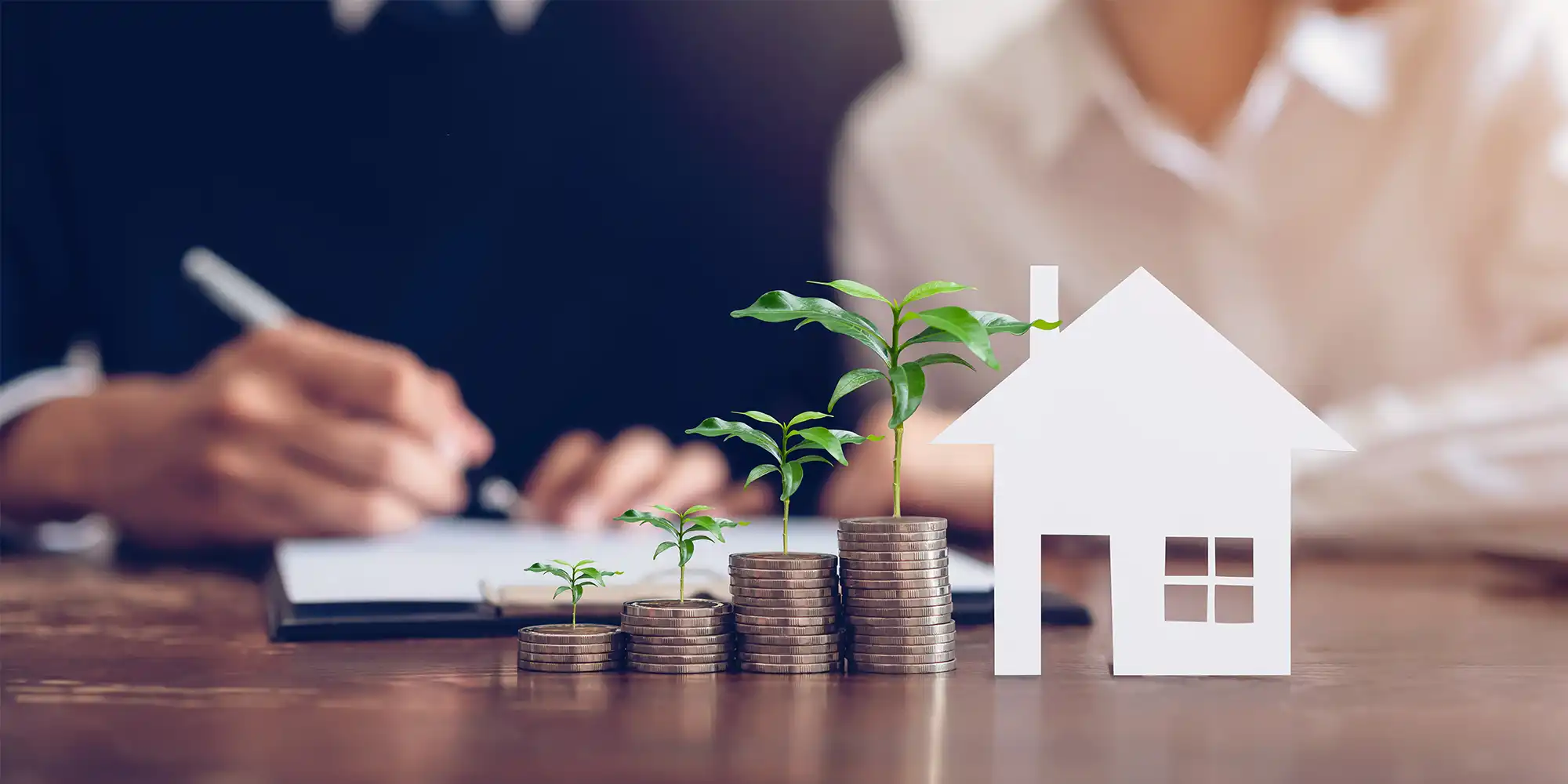 caucasian man and woman signing papers with little white house and coin stacks sprouting plants in front