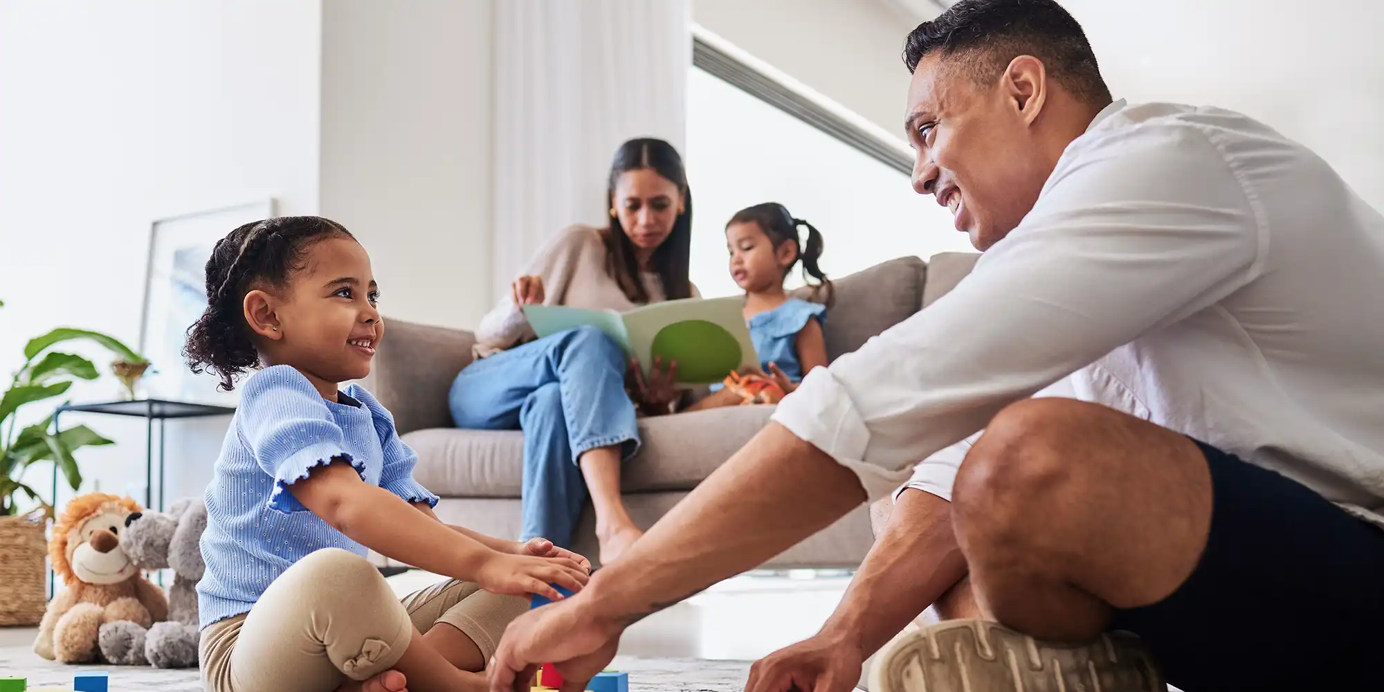 young BIPOC family of dad mom and two daughters reading books and playing blocks in bright modern livingroom
