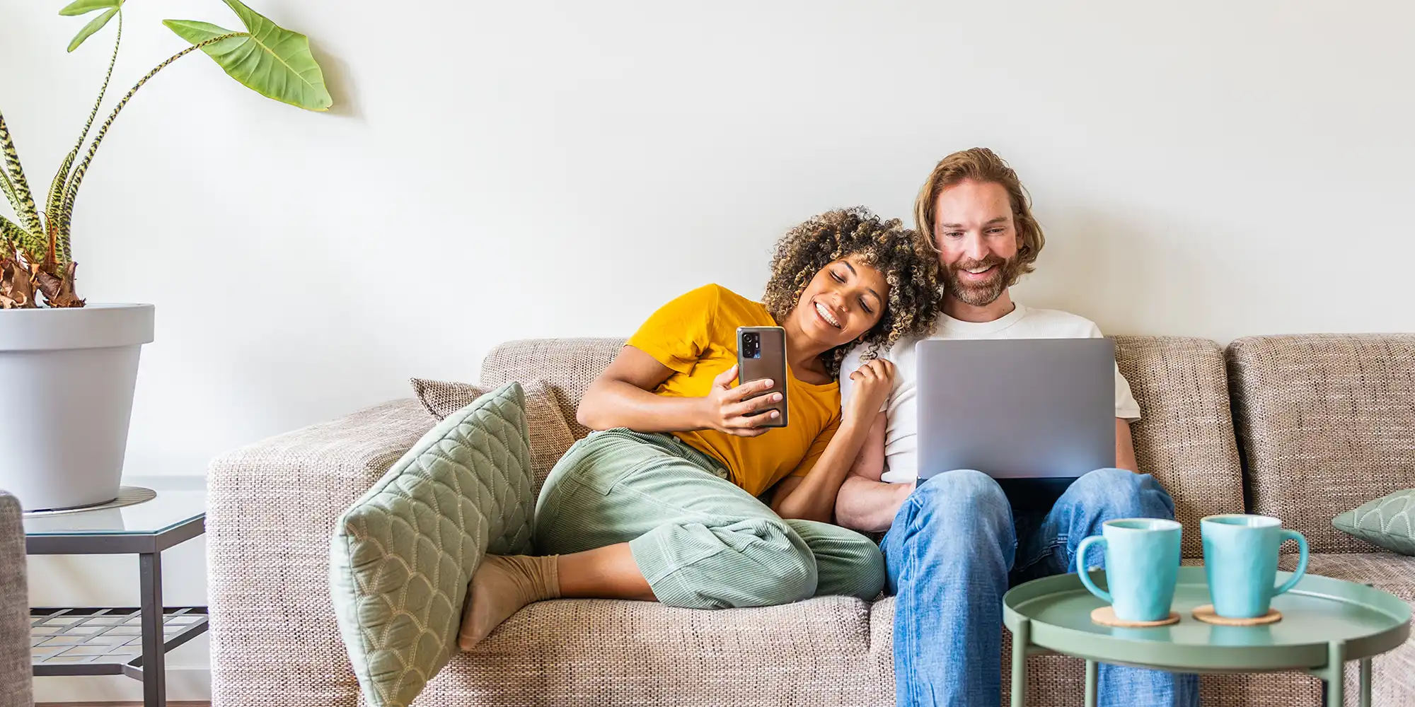 BIPOC young woman with curly multi-tone hair and caucasian man with red hair sitting on beige couch looking at laptop in modern livingroom