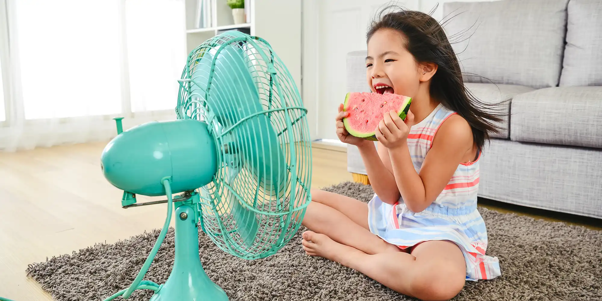 elementary age asian american girl in a striped dress eating large slice of watermelon while sitting in front of teal colored fan blowing air