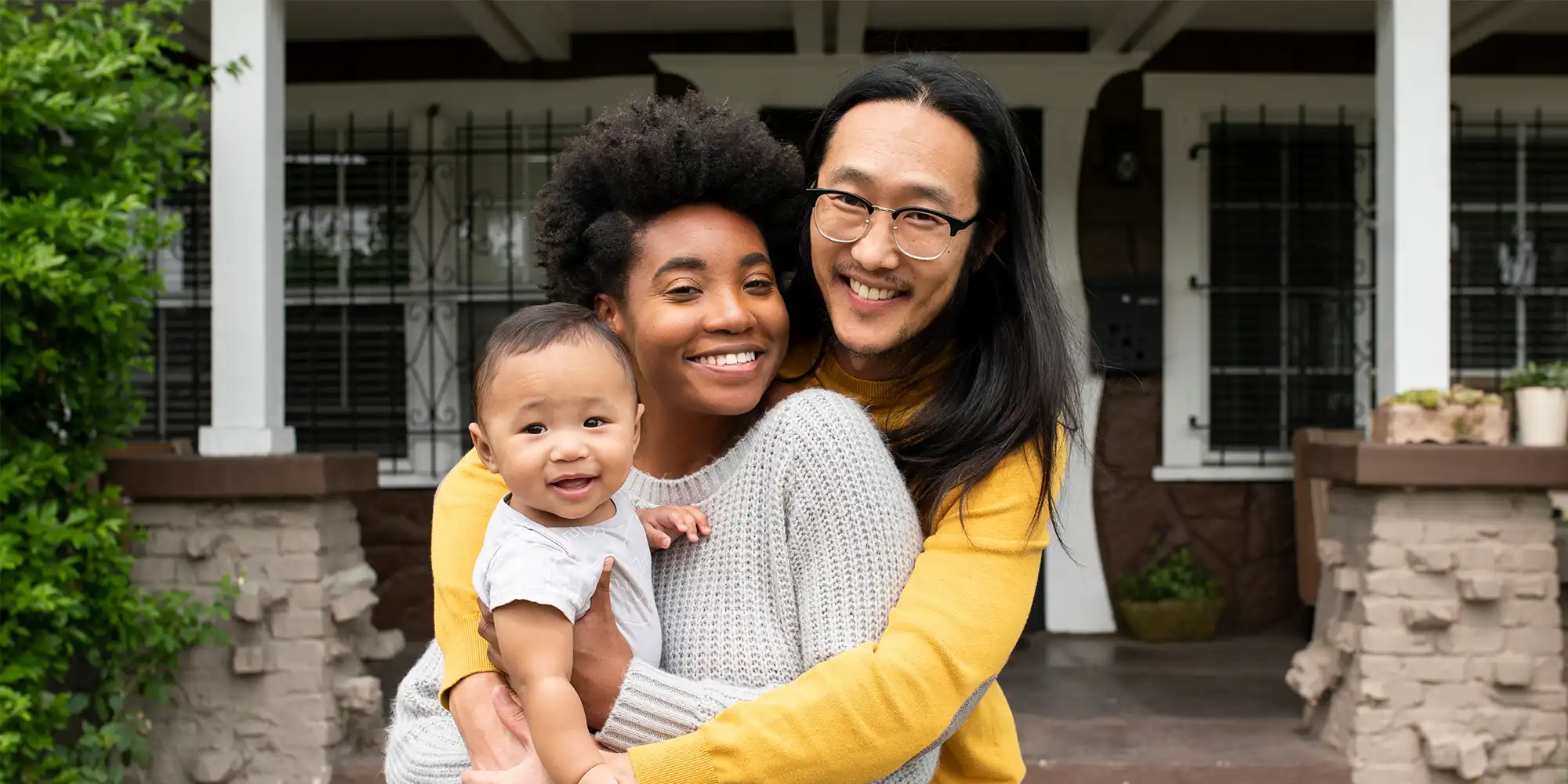 asian american man and african american woman and their baby embracing while smiling in front of their craftsman style home