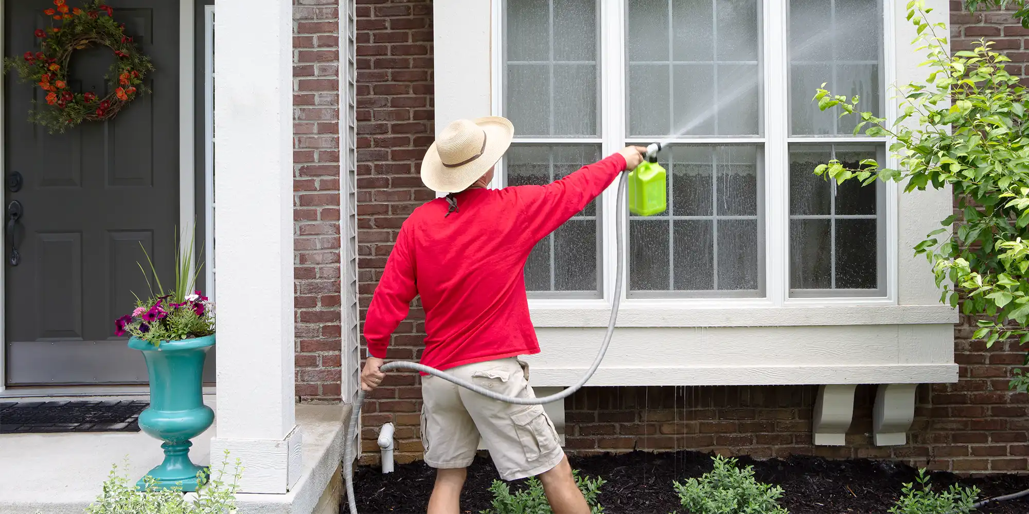 back view of woman wearing red shirt and khaki shorts washing windows outside with garden hose
