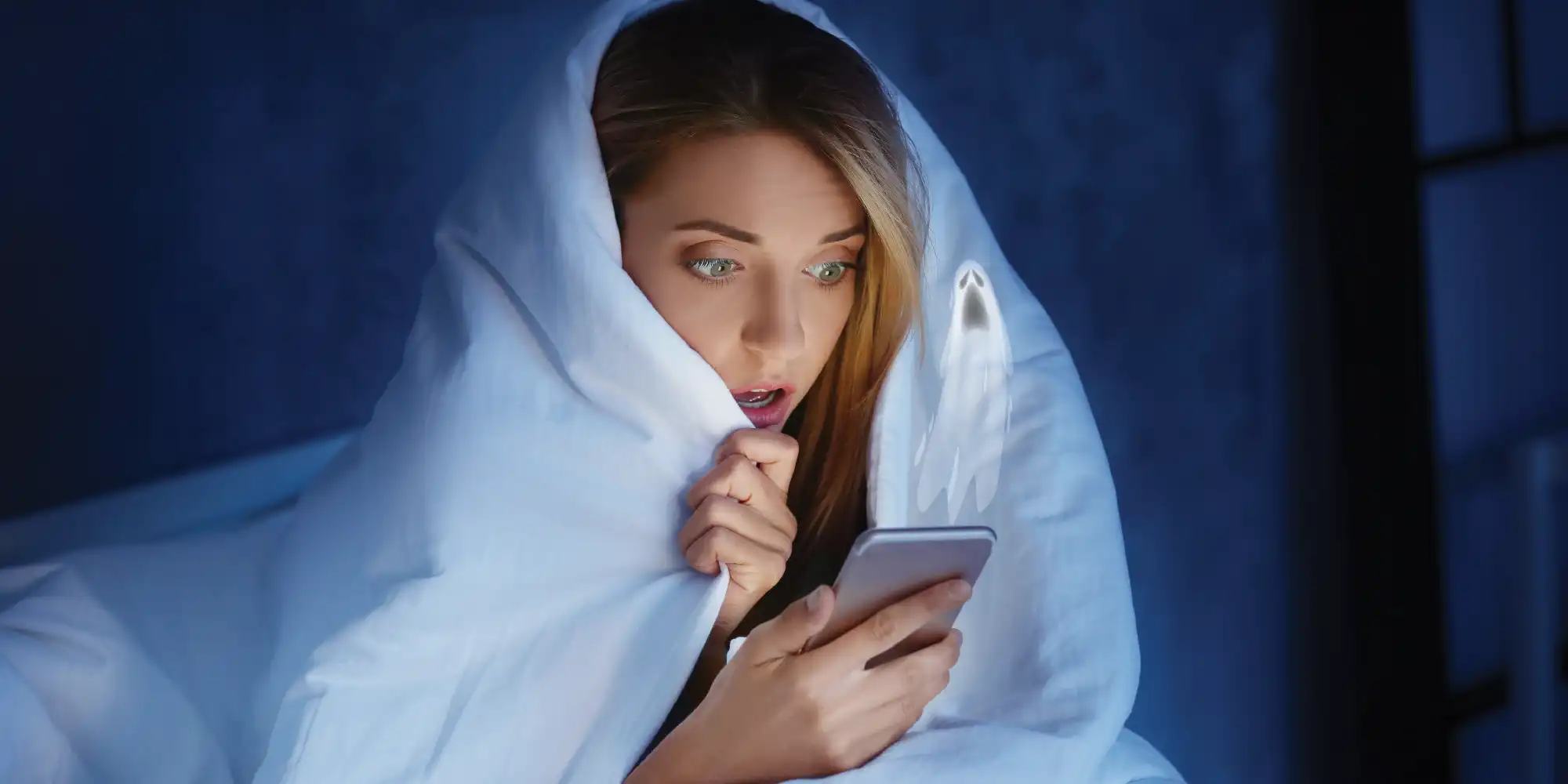 caucasian young woman with blond hair sitting wrapped in white blanket looking at ghostly phone with worried expression