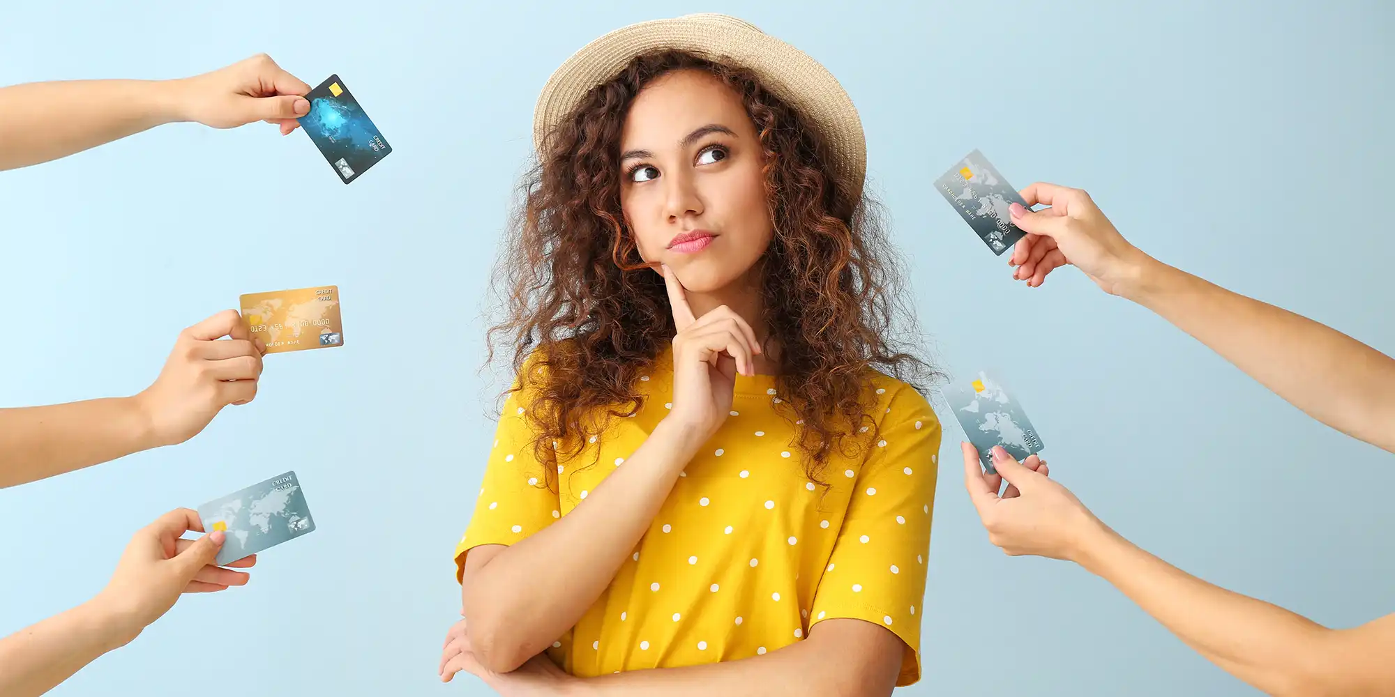young adult woman with curly brown hair in yellow top pondering the credit cards surrounding her