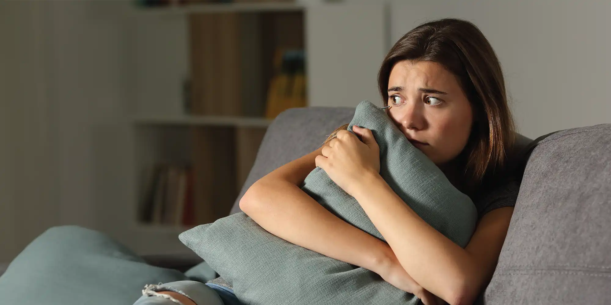 young caucasian woman with medium length brown hair looking scared while clutching a sage color pillow while sitting on a grey couch