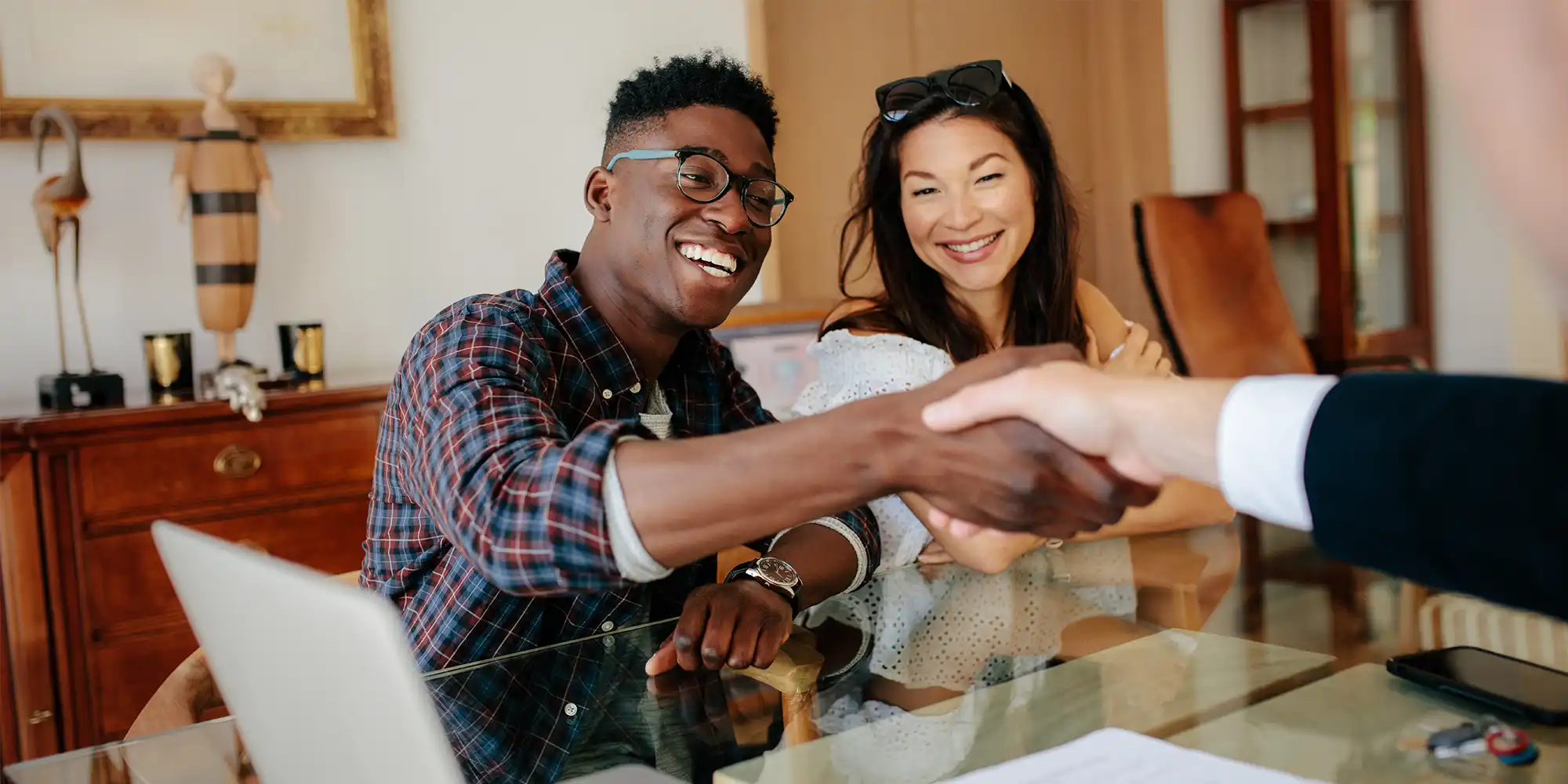 African American man wearing glasses is shaking the hand of someone across a desk while sitting next to a woman with dark hair