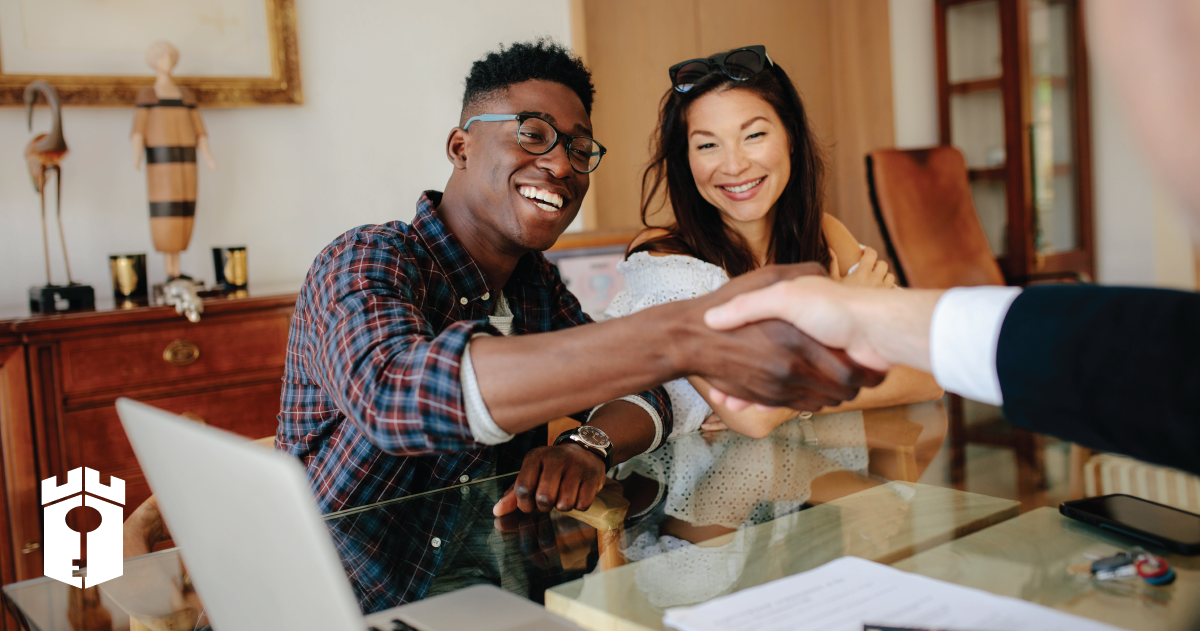 African American man wearing glasses is shaking the hand of someone across a desk while sitting next to a woman with dark hair